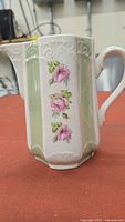 Front view of the vintage porcelain creamer showing multiple pink rose blossoms with green leaves on embossed white porcelain with green panel accents.