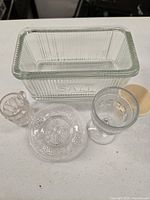 Clear glass ribbed Hoosier salt box with lid alongside three additional glassware pieces on a white surface.