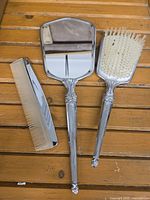 Full view of the silver plated vanity set: hand mirror, hairbrush, and comb laid on wooden surface.