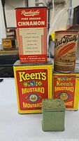Photo showing four vintage tins: two yellow Keen's Mustard tins, one red and white Rawleigh's Pure Ground Cinnamon tin, and one yellow and brown cylindrical Red Feather Baking Powder tin, with a small green spice tin in front.