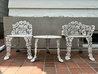 Front view of two Victorian cast iron grape vine chairs and round patio table on brick floor with beige wall background.