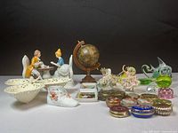 Full lot showing porcelain colonial couple figurine, globe, glass animal figurines, trinket boxes, and other decorative items on a white surface against black background.