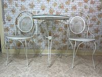 Set of white metal bistro table and two chairs against floral wallpaper, showing overall condition and design.