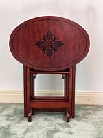 Front view of four oval wooden TV trays stacked upright on their folding wooden stand. Decorative black inlay pattern visible on front tray surface.