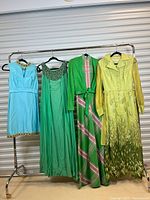 Front view of four dresses hanging on rack: blue sleeveless, green sleeveless, green/pink plaid with jacket, yellow long sleeve embroidered.