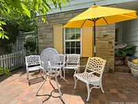 Full view of white metal patio set including round table, four chairs, yellow umbrella in stand on brick patio.