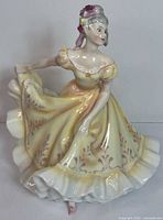 Front and partial side view of Royal Doulton Ninette figurine, showing the detailed front of the yellow dress and the figure's pose.