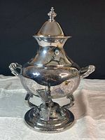 Front angled view of the silver plated coffee urn showing its body, handles, hinged lid with finial, and pedestal base.