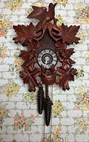 Front view of hand-carved cuckoo clock with pinecone weights against floral wallpaper background