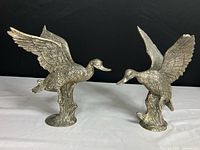 Pair of silver-colored metal duck statues facing each other with wings raised, placed on a white surface against black background.