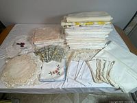 Full overview of assorted vintage table linens laid out on a white covered table showing stacks of folded linens including various placemats, doilies, napkins and tablecloths