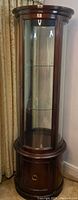 Full view of round vintage curio cabinet showing curved glass front, two glass shelves, and wooden base storage door.