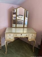 Front view of vintage cream-colored vanity with attached tri-fold mirror in gold frames, showing top, legs, and drawers.