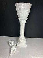 Front view of the white porcelain flower pot, pedestal, and cherub wall vase against black background.