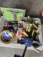 Photo of a container with various clamps including blue quick-release bar clamps, yellow and black spring clamps, coiled metal hose or cable, packaged hardware, and measuring tape.