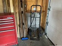 Black metal hand truck dolly with blue synthetic cleaning brush in a garage next to a red toolbox and garage door.
