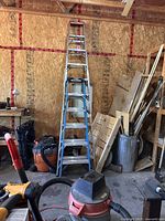Tall aluminum extension ladder leaning against wooden wall in workshop flanked by shop vacuum and tools