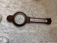 Wooden wall hanging barometer with round dial indicating barometric pressure and an attached thermometer, placed on carpeted floor.