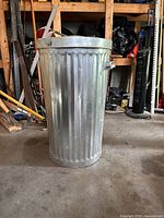 Front view of a tall galvanized metal garbage bin with ribbed vertical lines and two side handles in a workshop or garage setting.