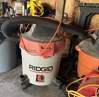 Ridgid shop vacuum cleaner with red and white body, black hose connected on top, sitting on a wheeled base in a workshop environment.