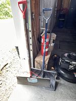 Steel snow shovel with blue handle and reinforced metal blade leaning against wooden wall inside garage or shed.