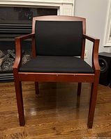 Front view of cherry red wooden framed accent chair with black fabric cushioned seat and backrest on wood floor, fireplace in background.