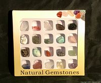 Wide view of the assortment of 24 natural gemstones arranged in a plastic grid within a labeled cardboard box, showing diverse colors and shapes.