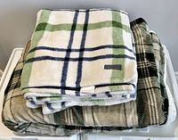 Two folded fleece blankets stacked, one white with green and navy plaid, one beige with black plaid, both soft fleece