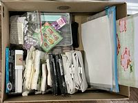 Top view of the boxed lot showing assorted cross stitch materials including canvas pieces, floss bundles, needles, and storage pouches.