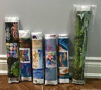 Photo showing 6 rolled diamond art kits standing on floor against a wall. Each roll has a preview image of the design and printed codes for diamond placement on canvas.