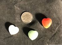 Three polished heart stones (white quartz, green aventurine, red agate) placed next to a dime for size reference on a dark fabric background.
