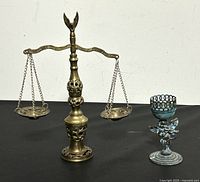 Photo showing both vintage brass items: balance scale and candle holder on a black surface.