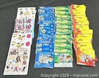 Top-down image showing 30 assorted sticker packages arranged in rows by color and design, including blue, green, and yellow packaged stickers with cartoon and Disney themes.