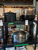 Black speckled enamel stockpot and food dehydrator on top shelf, large stainless steel roasting pan on bottom shelf of black metal shelving unit.