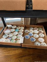 Two boxes containing numerous small vintage butter pat plates with varied floral and geometric decorative patterns.