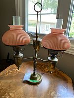 Photo showing entire double arm table lamp positioned on a wooden table by a window. Two pink ribbed glass shades with clear cylindrical tops mounted on brass arms. Lamp base is round and brass with a central post and a ring-shaped handle at the top.
