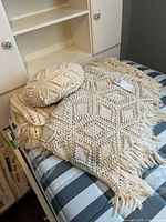 Set of three items, round crocheted pillow on top of two folded crocheted afghan blankets with textured geometric patterns and fringes in ivory cotton yarn.