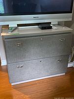 Front view of the large two drawer file cabinet with fabric drawer fronts and metal handles, drawer locks visible, cabinet topped with a large TV and small electronics.