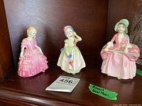 Three Royal Doulton figurines displayed on a shelf: Rose in pink dress, Babie holding umbrella, Bo-Peep seated with crook.