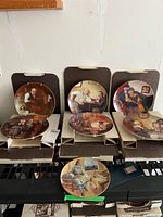 Set of seven Norman Rockwell collector plates displayed with original cardboard boxes. Plates depict different colorful scenes with gold rims.