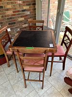 Full view of vintage wooden card table with black vinyl top and four Ferguson wooden chairs with pink vinyl seats arranged around the table on tile floor near brick wall and glass doors.