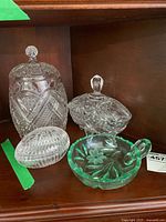 Four vintage cut glass pieces on wooden shelf: three clear glass covered dishes and one green glass handled dish