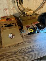 Wide view of vintage record collection including boxes and loose records on a wooden floor in a basement setting, some records with visible 45 RPM and 78 RPM formats.