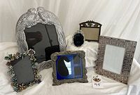 Six decorative picture frames arranged in two rows; includes a large lovebirds pewter frame, a bronze geometric frame, floral rhinestone frame, and others.