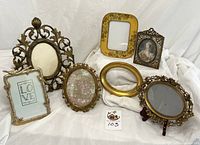 Seven tabletop vintage and vintage-style picture frames arranged on white cloth, showing various ornate designs including floral, rococo, and ribbon motifs. Includes gilt, brass, and gold finishes.