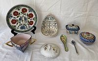 Overview of colorful assorted pottery lot with various plates, bowls, spoons, and covered dishes arranged on white cloth background.