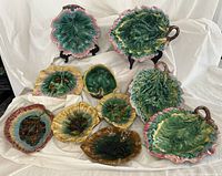 All ten leaf motif majolica plates and dishes arranged on cloth. Four handled serving plates at back, six smaller dishes front.