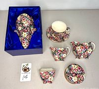 Seven pieces of Royal Winton porcelain items with floral pattern on black background. Wall pocket in original box shown alongside teacup with saucer, creamer, sugar bowl, and small vase.