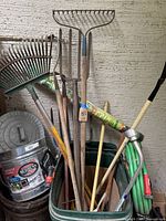 Green rolling bin containing assorted garden tools including rakes, hoes, a metal hose sprayer, green coiled garden hose, and galvanized steel container.