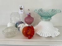 Overall collection of hobnail glass items shown on mantel: includes white ruffle bowl, mermaid glow bowl, two bud vases, lidded jar, pitcher.